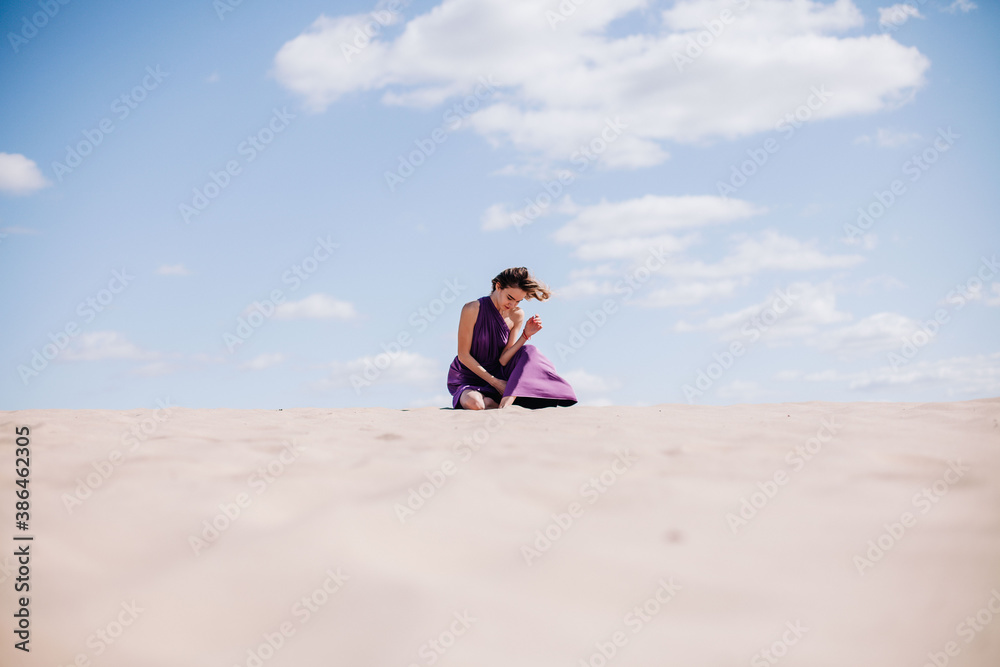 A young, slender girl in a beige dress with purple cloth in her hands posing in the desert in the wind