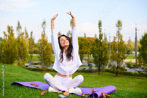 Attractive brunette girl in white sportwear sit on purple mat in lotus pose and stretch up hands under head, outdoor fitness, yoga exercises.