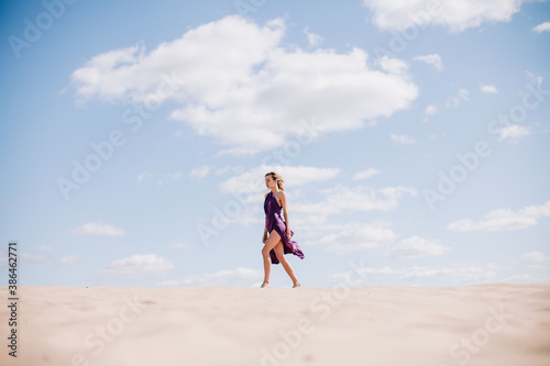 A young, slender girl in a beige dress with purple cloth in her hands posing in the desert in the wind © Denis