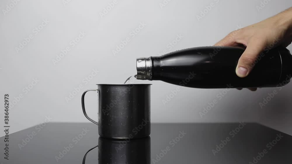 Close-up of male hand pouring water into the steel cup from stainless thermo bottle on black table of glass. Background of white color.