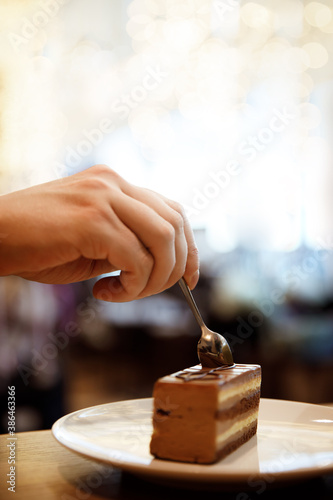 A man's hand breaks off a brown chocolate cake with a spoon. Dessert decorated with the musical symbol treble clef and a coffee drink in a glass on the table in a cafe. Copy spase. Selective focus.