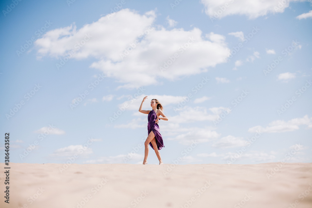 A young, slender girl in a beige dress with purple cloth in her hands posing in the desert in the wind