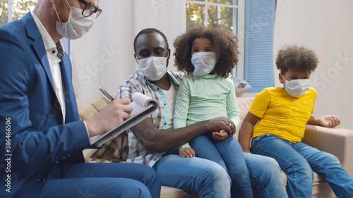Insurance agent in mask consulting smiling young african man with kids at home.
