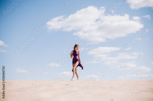 A young, slender girl in a beige dress with purple cloth in her hands posing in the desert in the wind