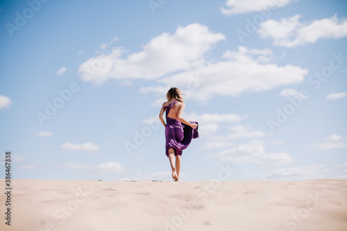 A young, slender girl in a beige dress with purple cloth in her hands posing in the desert in the wind