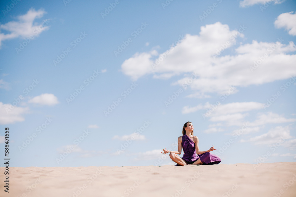 A young, slender girl in a beige dress with purple cloth in her hands posing in the desert in the wind