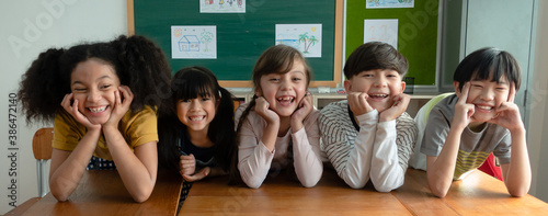 Portrait of happy elementary international students sitting on desk touches cheeks with both hands and looking at camera in classroom.Web banner panorama with adorable school kids.Education concept.