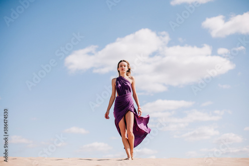 A young, slender girl in a beige dress with purple cloth in her hands posing in the desert in the wind