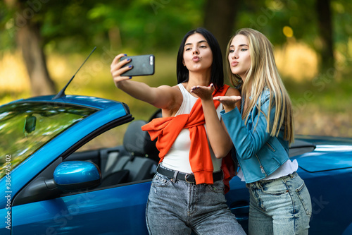 Young two women blow kiss taking a selfie while standing near convertible car on the street