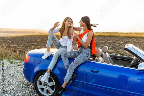 Young two women sitting on convertible car and talking spend free time together.