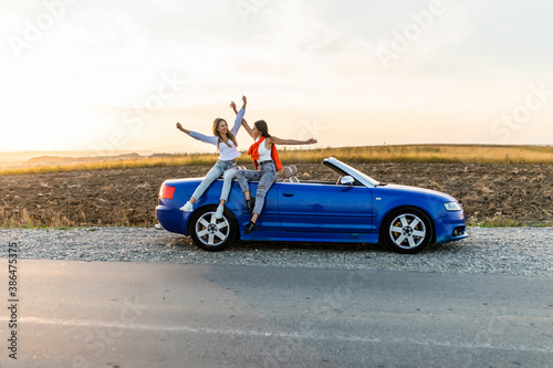 Fototapeta Naklejka Na Ścianę i Meble -  Couple of best women friends having fun together smiling while sitting on convertible car on sunset