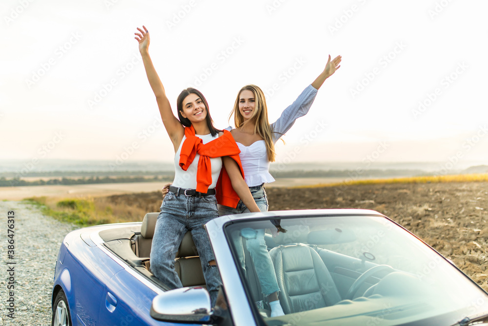 Fototapeta premium Happy two women in car driving on road trip. Young two women having fun dancing and cheering in car driving on travel vacation together.