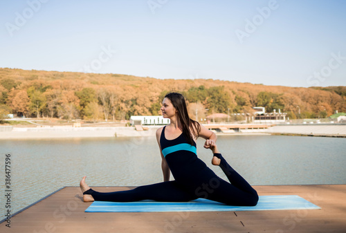the athlete performs stretching on a blue Mat and looks away on the pier against the background of the lake