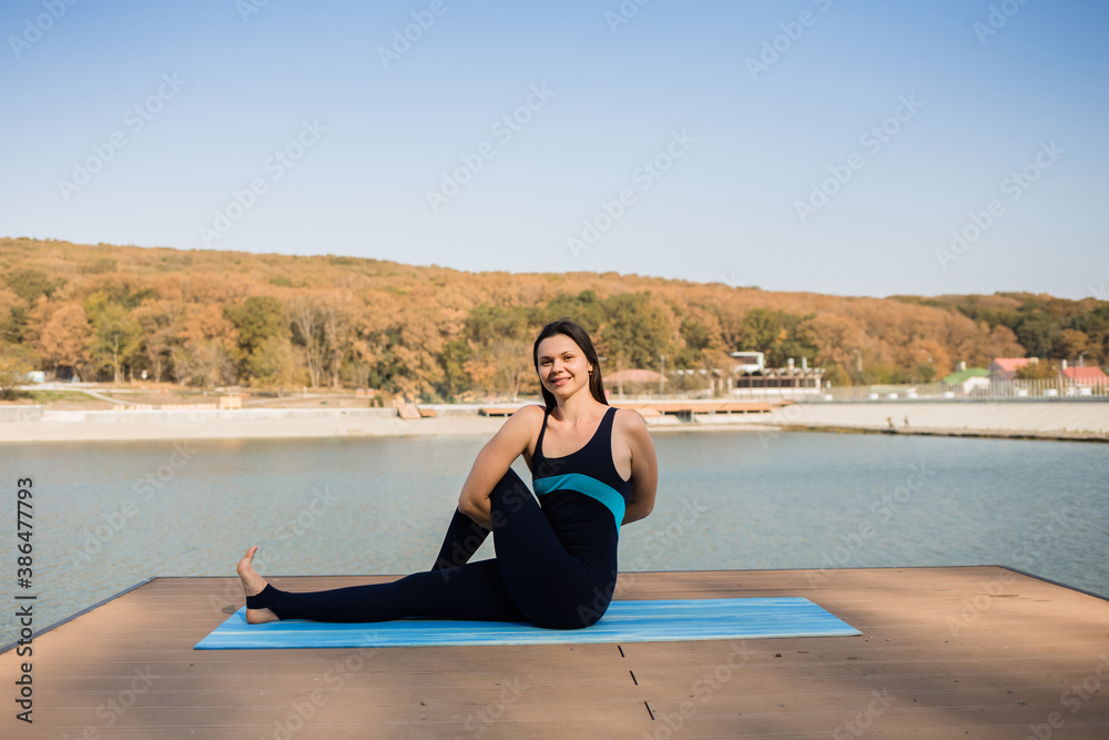 a female yogi performs the Marichiasana asana on a blue Mat on a pier with a lake background