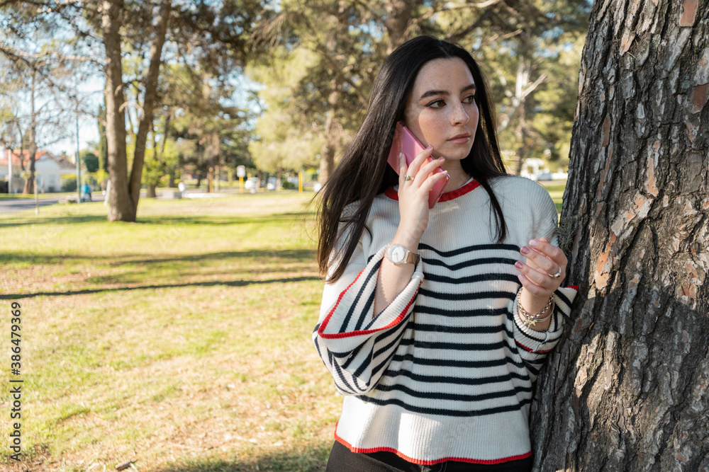 Black-haired girl with freckles using her phone next to a tree outdoors. Black-haired girl with freckles using her phone next to a tree outdoors.