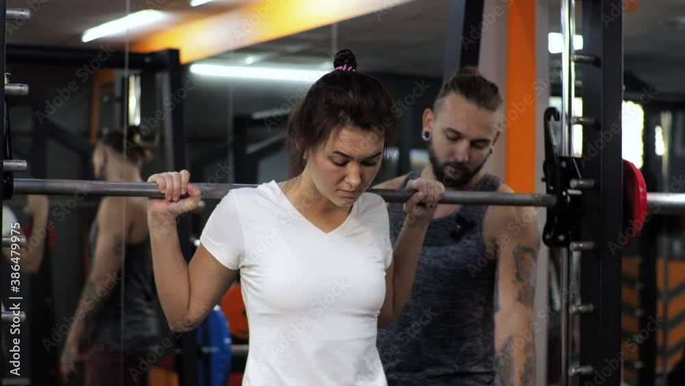 Gym. A funny man is training an aspiring young woman. Teaches you to swing your legs with squats with a barbell. Motivates to exercise. Gym. A funny man is training an aspiring young woman. Teaches you to swing your legs with squats with a barbell. Motivates to exercise.