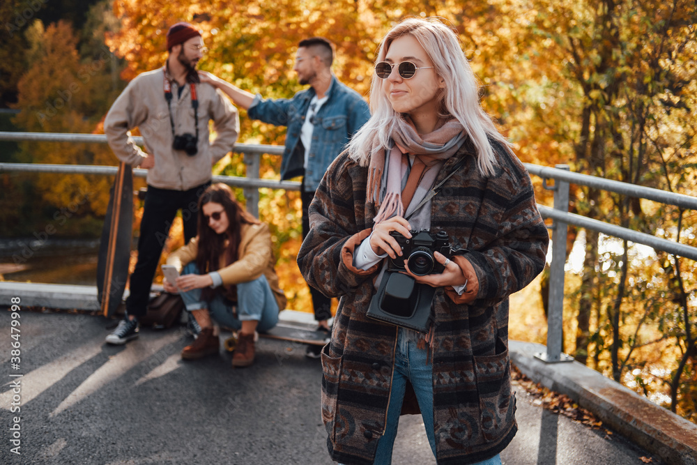 Obraz premium Pretty girl with blond hairs and camera posing in foreground in autumn landscape background and her friends in daytime.