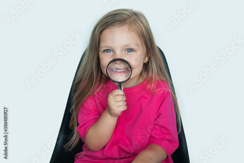 Blonde caucasian girl is using a magnifying glass on her opened mouth smiling at camera on a white studio wall photo