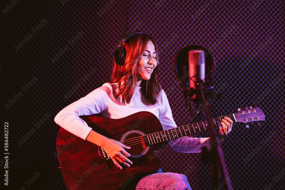 Obraz premium Pretty Asian female singer recording songs by using a studio microphone and pop shield on mic while playing guitar and wearing headphones in the studio. Performance and show in the music business.