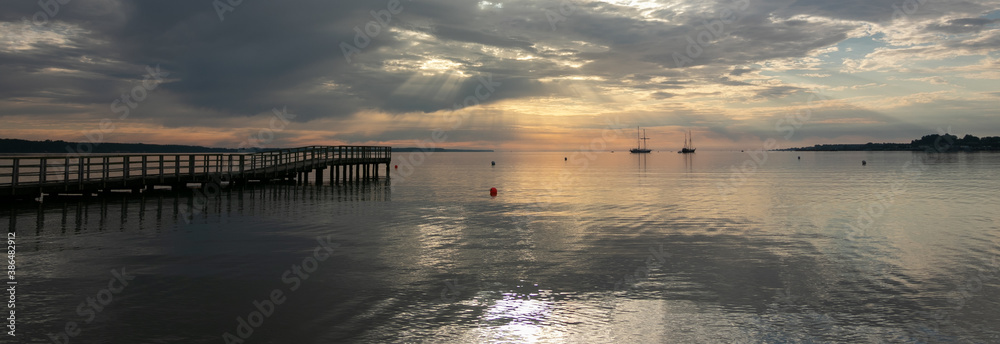 panoramic view to a tranquil sea scenery with jetty and sailing ship with dramatic sun rays
