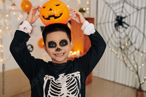 Portrait of funny boy in skeleton costume smiling and posing with jack o lantern on his head at Halloween party photo