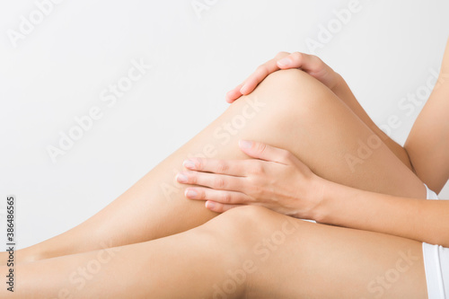 Young adult woman sitting and touching beautiful smooth legs. Isolated on light gray background. Daily care about clean and soft body skin after shaving or depilation. Closeup. Side view.
