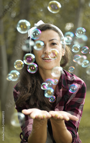 Brunette woman with a plaid jacket blowing some bubbles with trees in a blurry background