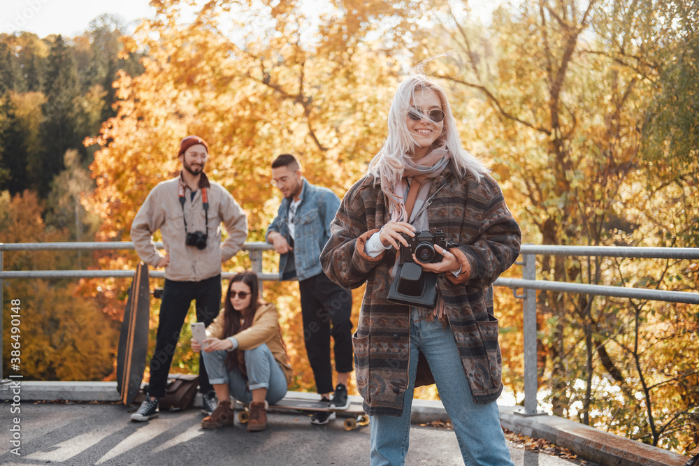 Obraz premium Pretty girl with blond hairs and camera posing in foreground in autumn landscape background and her friends in daytime.