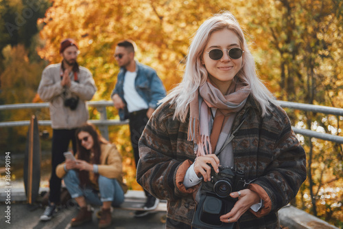 Fashionably dressed blond girl with sunglasses and camera having a good time with her friends in autumn public park.