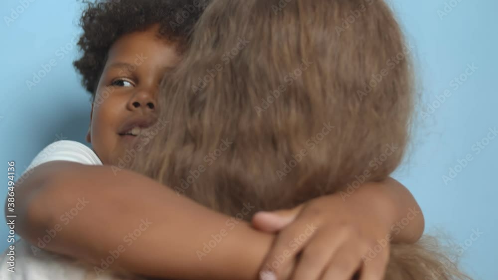 Close up of cute afro preteen boy embracing mother isolated on blue background Close up of cute afro preteen boy embracing mother isolated on blue background