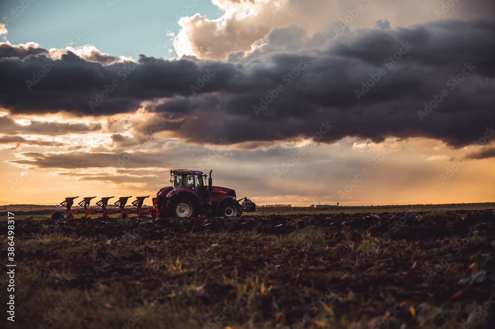 Fototapeta premium Tractor plowing the fields in the countryside by sunset. Agricultural tractor plowing the field. Red Tractor with plow. Sunset over the autumn field.