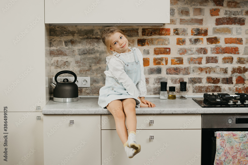 A happy young blonde girl with pale skin is posing while sitting on the table in the kitchen. A happy young blonde girl with pale skin is posing while sitting on the table in the kitchen.
