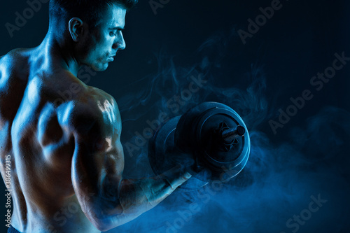 Muscular model sports young man with dumbbells in hand on dark background. Fashion portrait of strong brutal guy. Sexy torso. Male flexing his muscles.