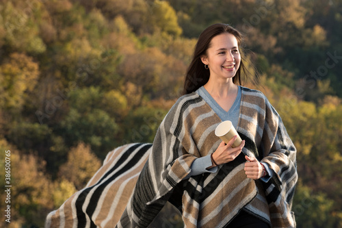 Beautiful moman in a poncho and a coffee in hand on nature background. Brunette girl smiling