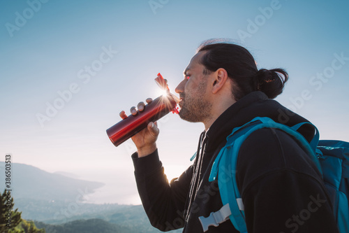 A man hiker drinks tea from a thermos on a mountain top at dawn.