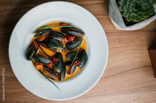 Gourmet serving of spicy tomato soup with fried mussels and chili peppers on a plate and wooden table isolated. Delicious seafood mussels . Lemon and baguette . Clams in the shells. Top view.