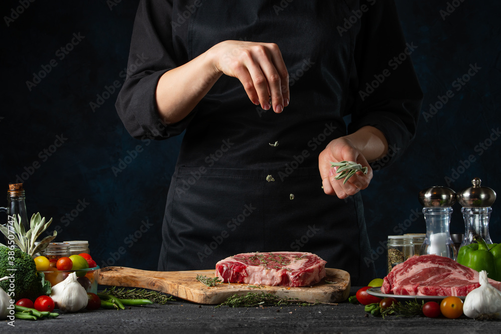 The chef in black uniform pours rosemary on steak on the wooden board on dark blue background. Preparing raw beef or pork. Backstage of cooking dinner. Frozen motion. Close-up view.