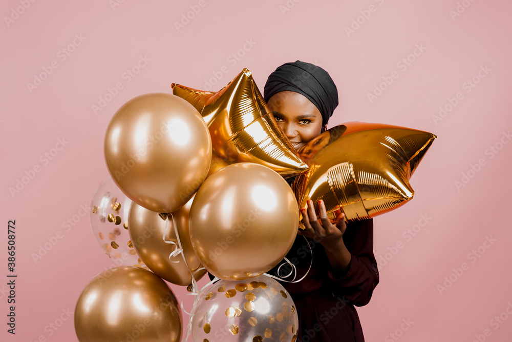 Black girl between golden ballons smiles and have a party isolated on blank pink background for advert . African woman celebrate holiday. Happy emotions of muslim young woman