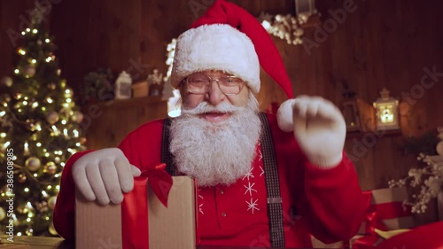 Happy Santa Claus, Saint Nicholas waving hand holding present talking to camera video calling kid or recording Merry Christmas virtual greetings on xmas eve sitting at table at home, webcam view.