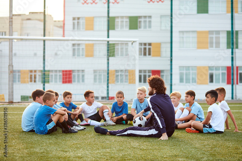 friendly kid boys have rest with trainer during football competition, confident male trainer give advice, explain and have talk with sportive boys, in stadium