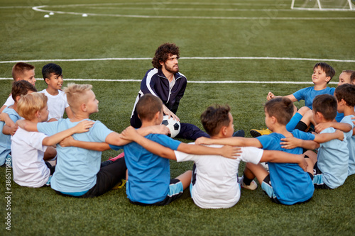 friendly kid boys have rest with trainer during football competition, confident male trainer give advice, explain and have talk with sportive boys, in stadium