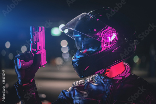 A cyberpunk biker concept. Motorbiker in a helmet with a toy gun is standing on a night city street in the neon lights.