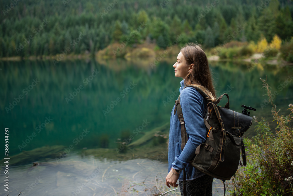 Young beautiful woman standing near amazing lake with clean blue water on wooded mountains background. Concept of friendly with nature. Close-up side view. Enjoyment of clear air. Relaxation. - obrazy, fototapety, plakaty 