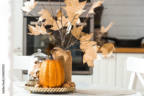 Branches with Golden leaves and a pumpkin on a tray. In the background-the interior of a white kitchen in the Scandinavian style. The concept of home comfort. Autumn decor for the Halloween holiday.