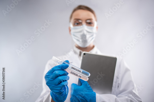 Doctor in white medical gown, protective blue gloves, mask and glasses keeps laptop and covid-19 vaccine on white background. Works moment. Healthcare and coronavirus infection concept.
