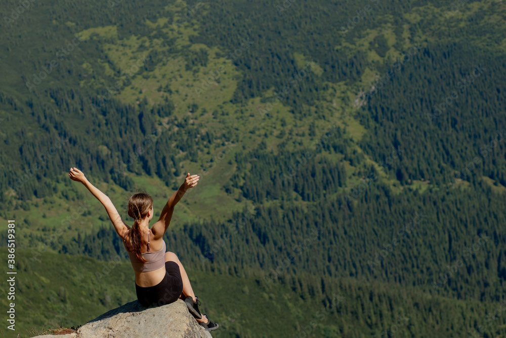 The young woman sitting at the top of the mountain raised her hands up on forest background. The woman climbed to the top and enjoyed her success.
