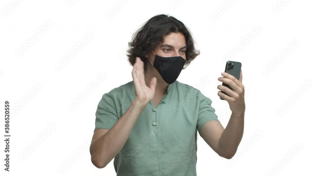 Coronavirus, social distancing and health concept. Hispanic guy in black mask having video call on smartphone, waving hand and saying hello at mobile phone, standing over white background Coronavirus, social distancing and health concept. Hispanic guy in black mask having video call on smartphone, waving hand and saying hello at mobile phone, standing over white background