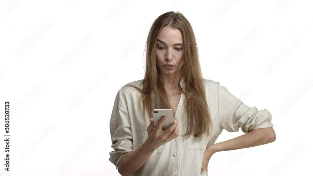Video shot of beautiful blond woman in casual blouse looking at mobile phone, checking messages and saying wow surprised, smiling pleased at camera, standing over white background Video shot of beautiful blond woman in casual blouse looking at mobile phone, checking messages and saying wow surprised, smiling pleased at camera, standing over white background