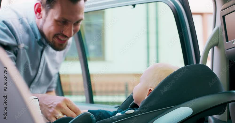 Closeup baby boy, who sits in the baby car seat inside of family car. Door opens and his smiled bearded father unfastens the safety belts, and carefully takes baby out of the car. Slow motion Closeup baby boy, who sits in the baby car seat inside of family car. Door opens and his smiled bearded father unfastens the safety belts, and carefully takes baby out of the car. Slow motion