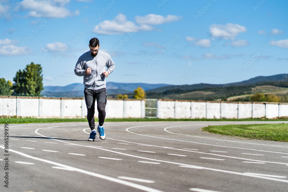 Full length front view on adult caucasian man jogging on the running track - Adult male athlete in stadium training in sunny summer or autumn day - real people healthy lifestyle concept copy space Full length front view on adult caucasian man jogging on the running track - Adult male athlete in stadium training in sunny summer or autumn day - real people healthy lifestyle concept copy space